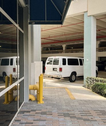 View of white vans parked in an urban multi-level garage, reflecting on glass facade.